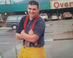 Standing in a parking lot, young man wearing red suspenders and yellow waterproof pants smiles. Parking lot is wet.