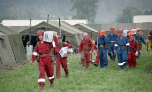 Hommes en combinaison rouge ou bleue portant des boîtes et des provisions. Tentes en arrière-plan.