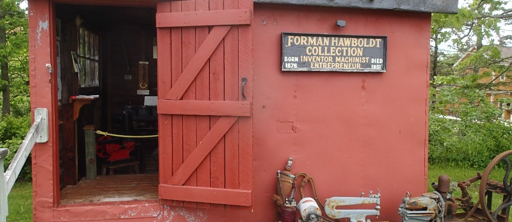 A small red metal building, with a doorway leading inside. A Hawboldt jet pump sitting in front with a sign that says Forman Hawboldt Collection.