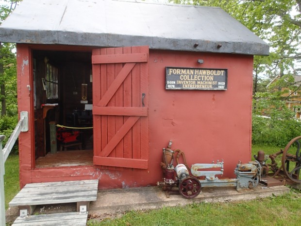A small red metal building, with a doorway leading inside. A Hawboldt jet pump sitting in front with a sign that says Forman Hawboldt Collection.