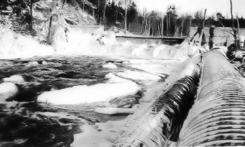 A black and white photo showing the dam on the East River with the gates closed, the water swirling at the foot of the dam and then continuing down the river. On the bank to the right, two large pipes that carried the water to the generating station.
