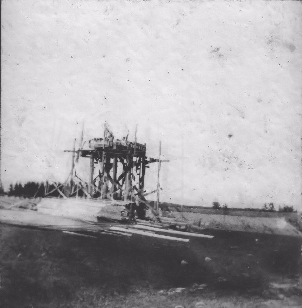 A black and white photograph showing the framework of a water tower being constructed with piles of lumber in the foreground and the embankment of the Halifax and Southwestern Railway bed in the background. This tower was to supply water for the village water supply and the trains.