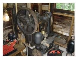 A large black iron pump with a flywheel. The water lines to and from the pump are shown as is a valve to turn off the water on the pipe. This was the pump used to supply the water throughout the summer to village residents. Currently it is on display at the Forman Hawboldt Collection building at Chester Train Station.