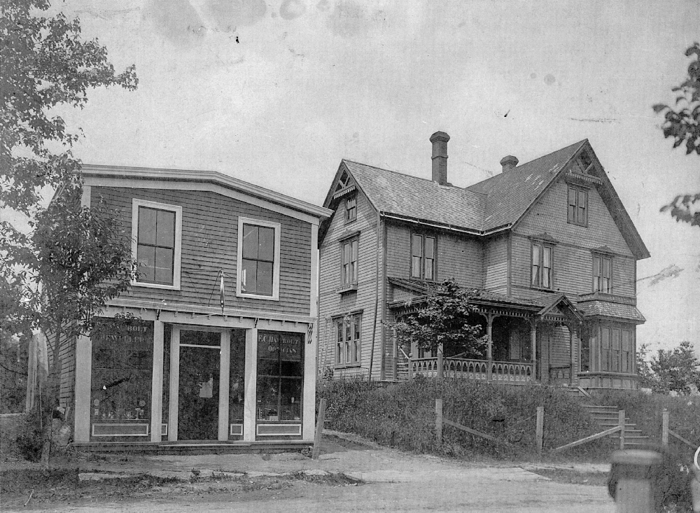 A black and white photo of the two-storey jewelry store with two shop windows and a door in the middle, close to the road. The top floor has two smaller windows. To the right on a rise, set back from the road, with a wire fence by the street, a three storey shingled house with steps leading up to the front door, dormer windows on the right of the front steps and a veranda along the left front of the house. The house sports gingerbread trim, many windows and a steeply pitched roof.