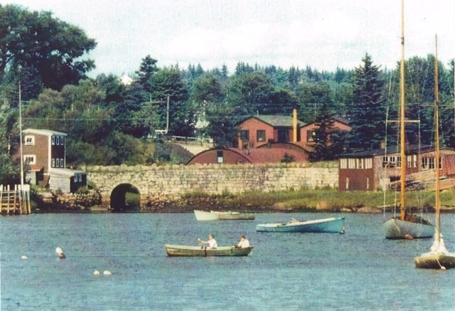 A colour photo with boats in the foreground looking from Mill Cove toward Hawboldt Gas Engines showing the old stone bridge, the red rounded foundry, machine shop and main office. A building on the left of the bridge was a storage building and the red buildings at the other end of the bridge were a woodworking shop. The stream came from Stanford Lake powering the foundry and supplying water for the village water system and empting into Mill Cove under the old stone bridge.
