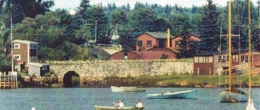 A colour photo with boats in the foreground looking from Mill Cove toward Hawboldt Gas Engines showing the old stone bridge, the red rounded foundry, machine shop and main office. A building on the left of the bridge was a storage building and the red buildings at the other end of the bridge were a woodworking shop. The stream came from Stanford Lake powering the foundry and supplying water for the village water system and empting into Mill Cove under the old stone bridge.