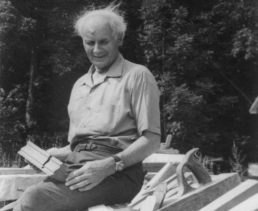 Black and white photograph of François-Xavier Lachance, aged, sitting outside on a pile of materials, holding a wooden molding.