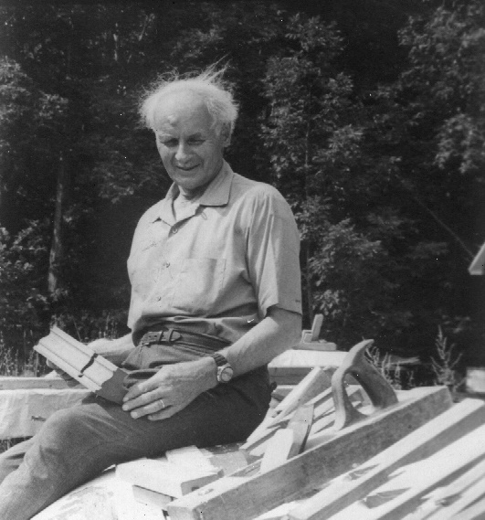 Black and white photograph of François-Xavier Lachance, aged, sitting outside on a pile of materials, holding a wooden molding.