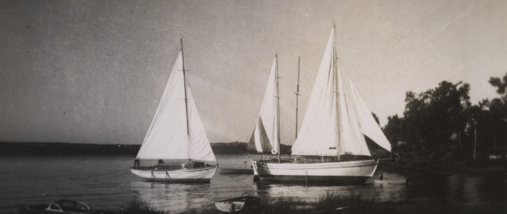 Black and white photograph of three white yachts, with hoisted sails, anchored near the shore.