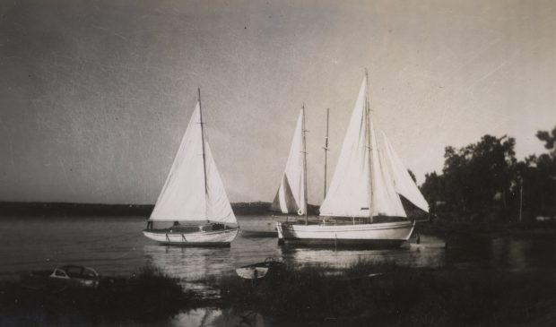 Black and white photograph of three white yachts, with hoisted sails, anchored near the shore.