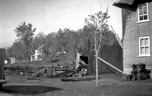 Black and white photograph showing the foundation of a building, an almost completely burned wall of wood and some burned planks on the ground. A stone residence, whose left wall and the roof of the house are blackened by fire, is partially visible to the right of the image.