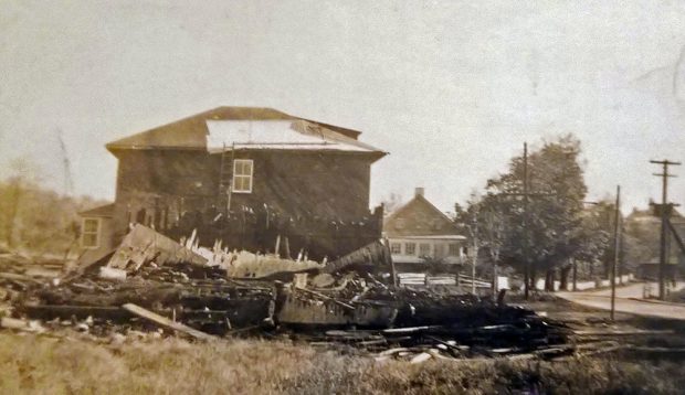 Sepia photograph showing a cluster of burned planks in the foreground. A two-storey residence damaged by fire is visible behind the remains.