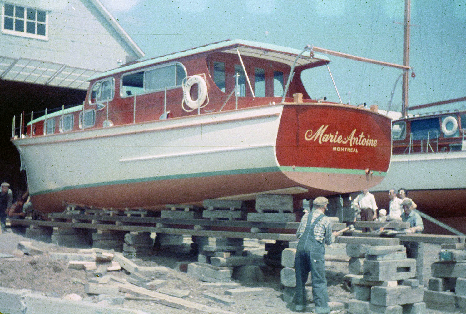 Color photograph where a white painted wooden yacht with mahogany cabin and aft is installed on blocks and boards at the exit of a hangar. The inscription "Marie-Antoine, MONTREAL" is at the back of the boat. F-X handles a hammer against the boards of the structure under the boat, helped by another man. Three adults and one child watch the scene from the side of the boat. A sailboat is partially visible in the background.
