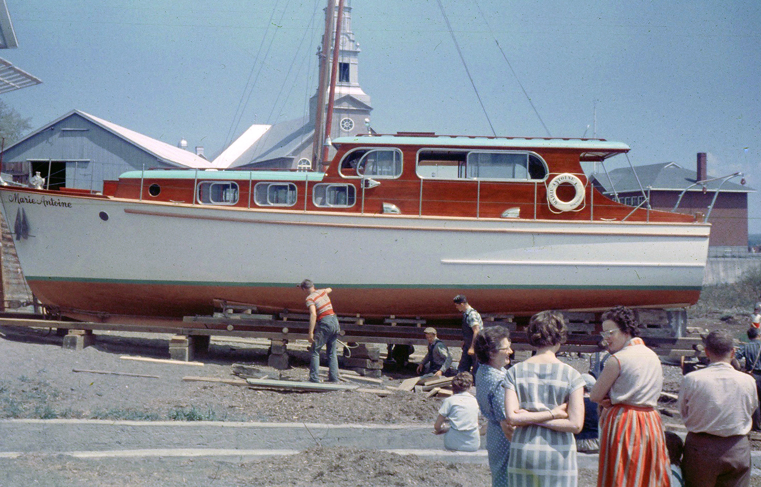 Color photograph where a wooden cruiser yacht painted in white, whose cabin is in mahogany, is installed on blocks and boards. The boat is shown in profile, with a church and a shed in the background. The inscription "Marie-Antoine" is at the front of the boat. Three men work on the structure under the boat. Three women discuss in the foreground. A man watches the scene.