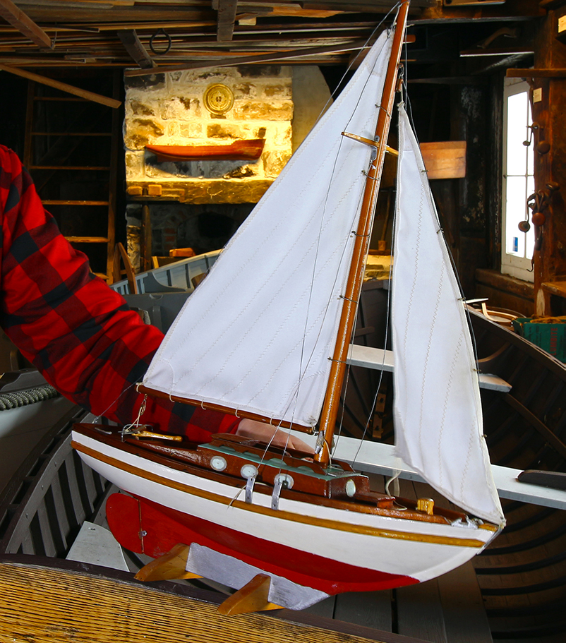 Color photograph of the model of a wooden sailboat, profile view. The boat is painted white, red and brown. The two white sails are hoisted.