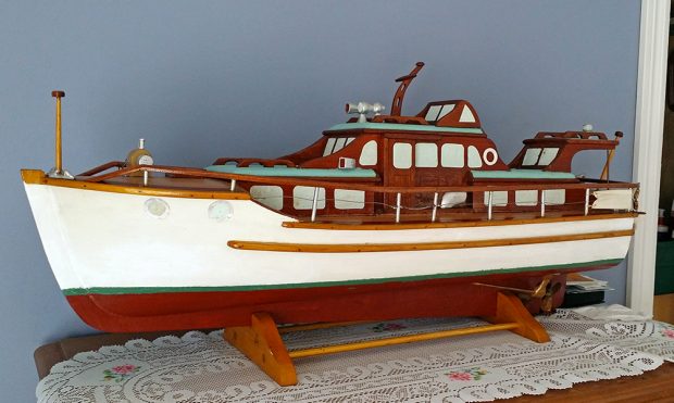 Color photograph of a model of a wooden yacht, profile view. The boat is painted white, red, green and brown. The object is exposed on a small wooden base, on the top of a piece of furniture.
