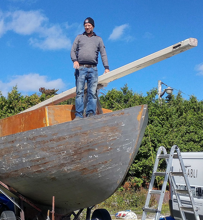 Low-angle color photograph showing the front of a gray painted wooden boat, on which stands a man (Jean-François Lachance), leaning on the lying mast of the boat on a trailer.