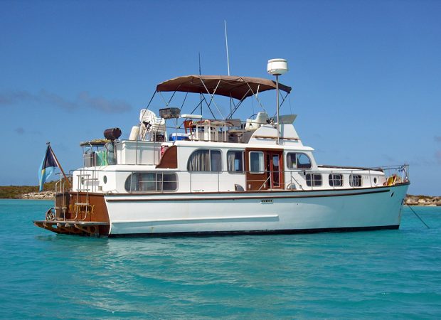 Color photograph of a white yacht, profile, on a blue sea. The upper deck is converted into a terrace with barbecue and canvas shelter.
