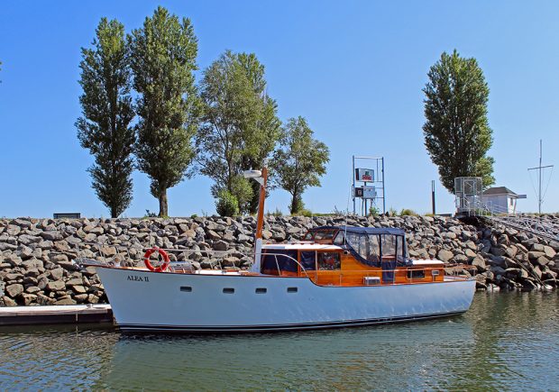 Color photograph of a white sailboat, profile, engraved ALEA II, moored to a wooden pontoon in front of a rocky breakwater. A pedestrian descent is on the right of the image.