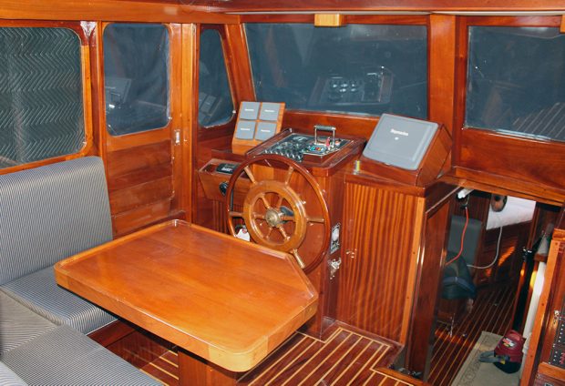 Color photograph of the interior of a cabin, the surfaces of which are mahogany woodwork. In the center is the wheel, in front of the navigation instruments. A wooden table partly surrounded by a bench is in the foreground.
