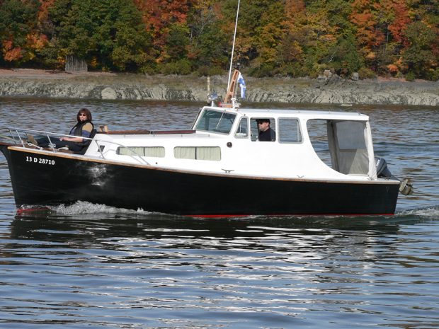 Color photograph of a small boat with black hull and white cabin, which sails in front of a rocky bank. A pilot is inside the cabin. A woman is sitting on the deck, at the front, smiling.