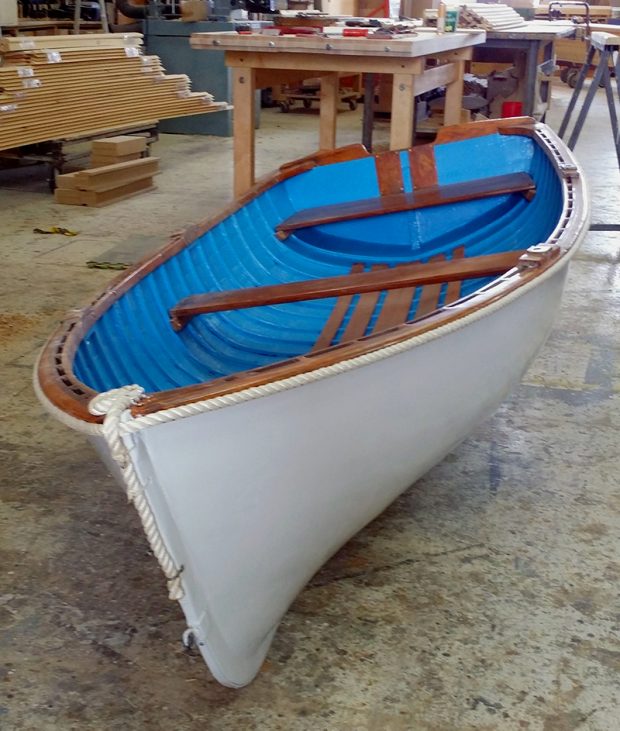Color photograph of a white wooden boat with blue interior, front view in light dive-angle. The boat is placed on a concrete floor in a carpentry workshop.