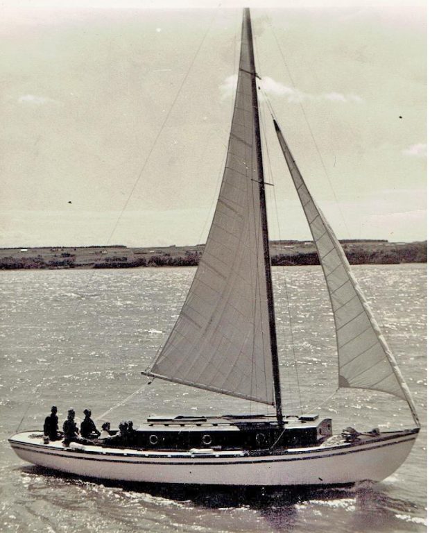 Photograph of a white sailboat with dark cabin, sailing sails deployed under the sun. Six people are on board. Fields are visible in the background.