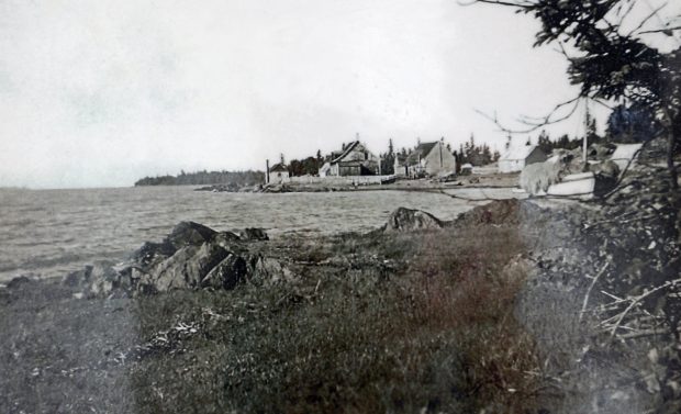 Black and white photograph showing four wooden buildings in the background. In front is a bay where a small masted boat is docked on the rocky shore.