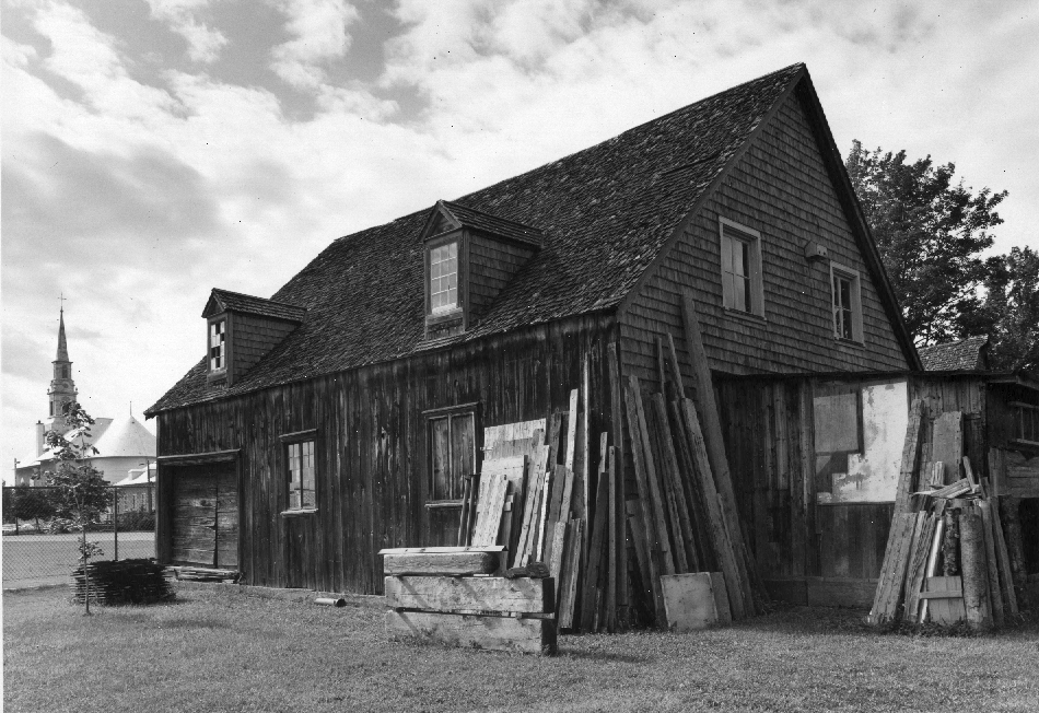 Black and white photograph of a wooden building and cedar shingles, with two dormers. Several boards and materials are backed on it. The church of Saint-Laurent is visible in the background, on the left.