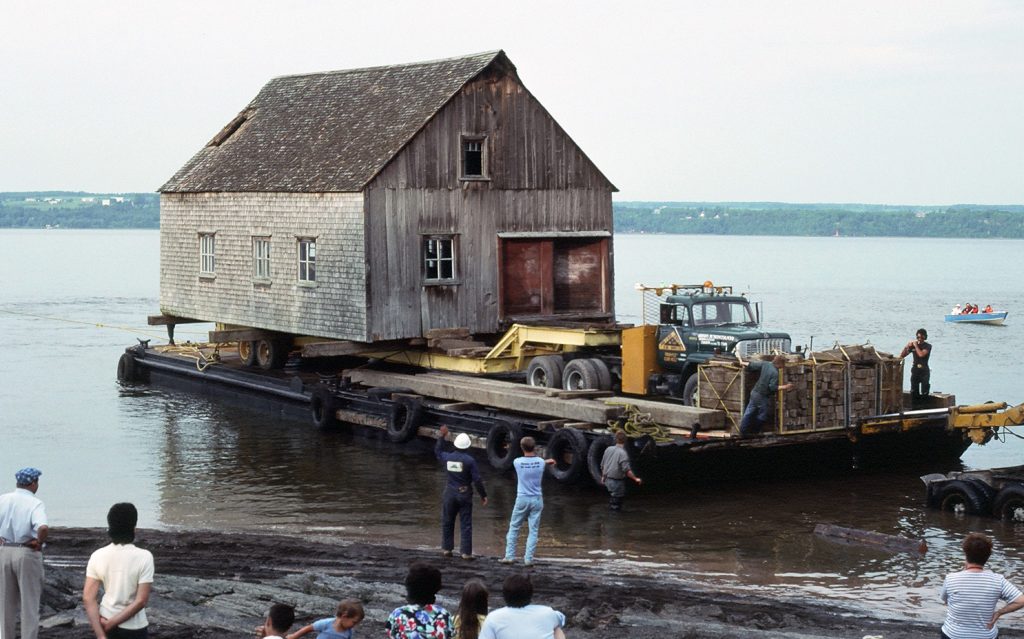 Photographie couleur montrant le déménagement par bateau d'une chalouperie en bois et bardeaux de cèdre. Le bâtiment est posé sur une remorque tirée par un camion, le tout monté sur une barge. Cinq travailleurs sont sur la barge ou près de la rive. Des curieux, enfants et adultes, observent la scène.