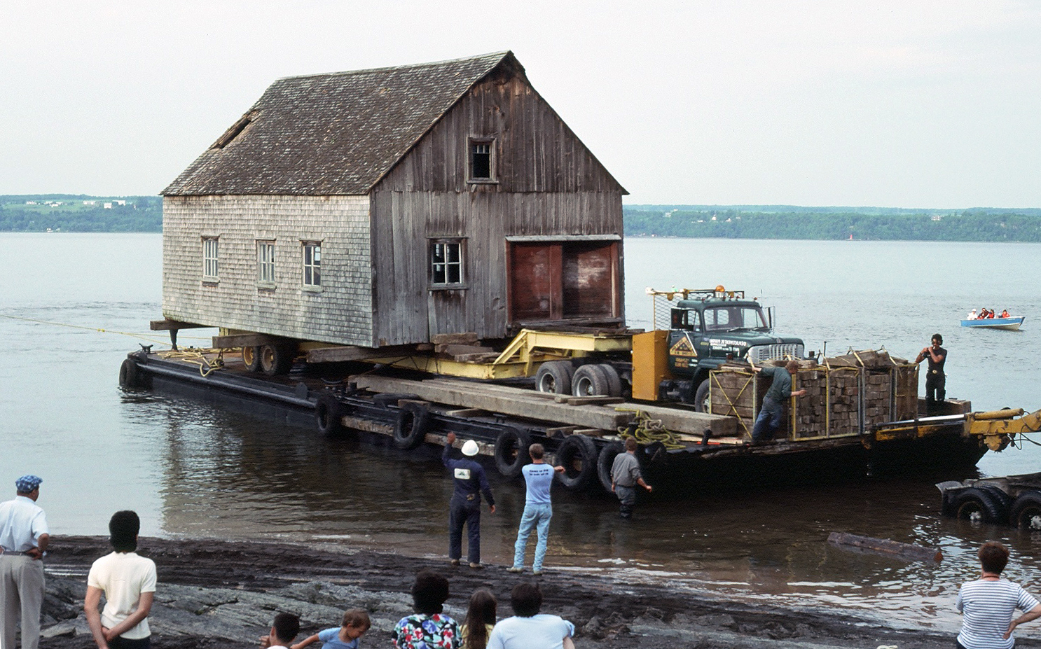 Color photograph showing the relocation by boat of a wooden and cedar shingles rowboat shop. The building sits on a trailer pulled by a truck, all mounted on a barge. Five workers are on the barge or near the shore. Curious children and adults watch the scene.