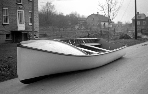 Black and white photograph of a large white wooden rowboat, looking brand new, on the road. Two houses, a field and a barn are visible in the background.