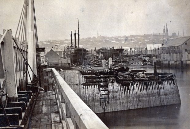 Black and white photograph of a shipyard. There is a dock in the foreground, as well as a sailboat with three masts and a second without mast in the center of the image. Large buildings are located on the water's edge on the right. The upper city of Quebec is visible in the background.