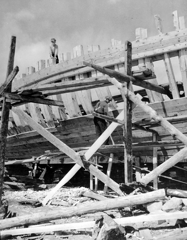 Black and white photograph of a man working to fix the siding of a large boat on pedestals. He is standing on a wooden scaffolding. A young boy is standing at the top of the hold structure.