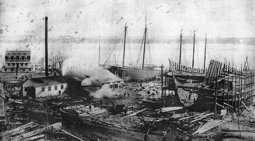 Black and white photograph of the Saint-Laurent shipyard. The structure of an large boat is on the right, in the foreground. Two sailboats under construction are in the center. The St. Lawrence River occupies the entire background, as well as the coast of the south shore.