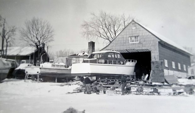Black and white photograph showing a cruiser yacht mounted on a base in front of a building with a large door, in winter. Four men are busy around the boat on the ground. Other boats are visible on the left.
