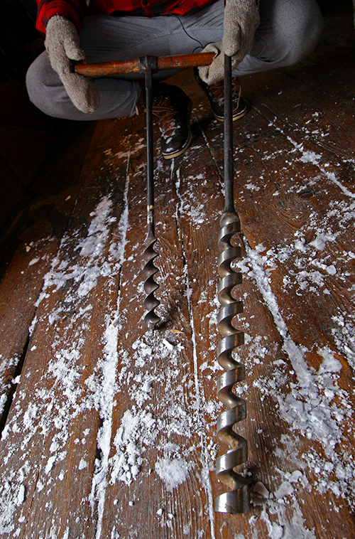 Color photograph of two large iron wicks, one of which has a wooden handle. The objects are held by Jean-François Lachance.