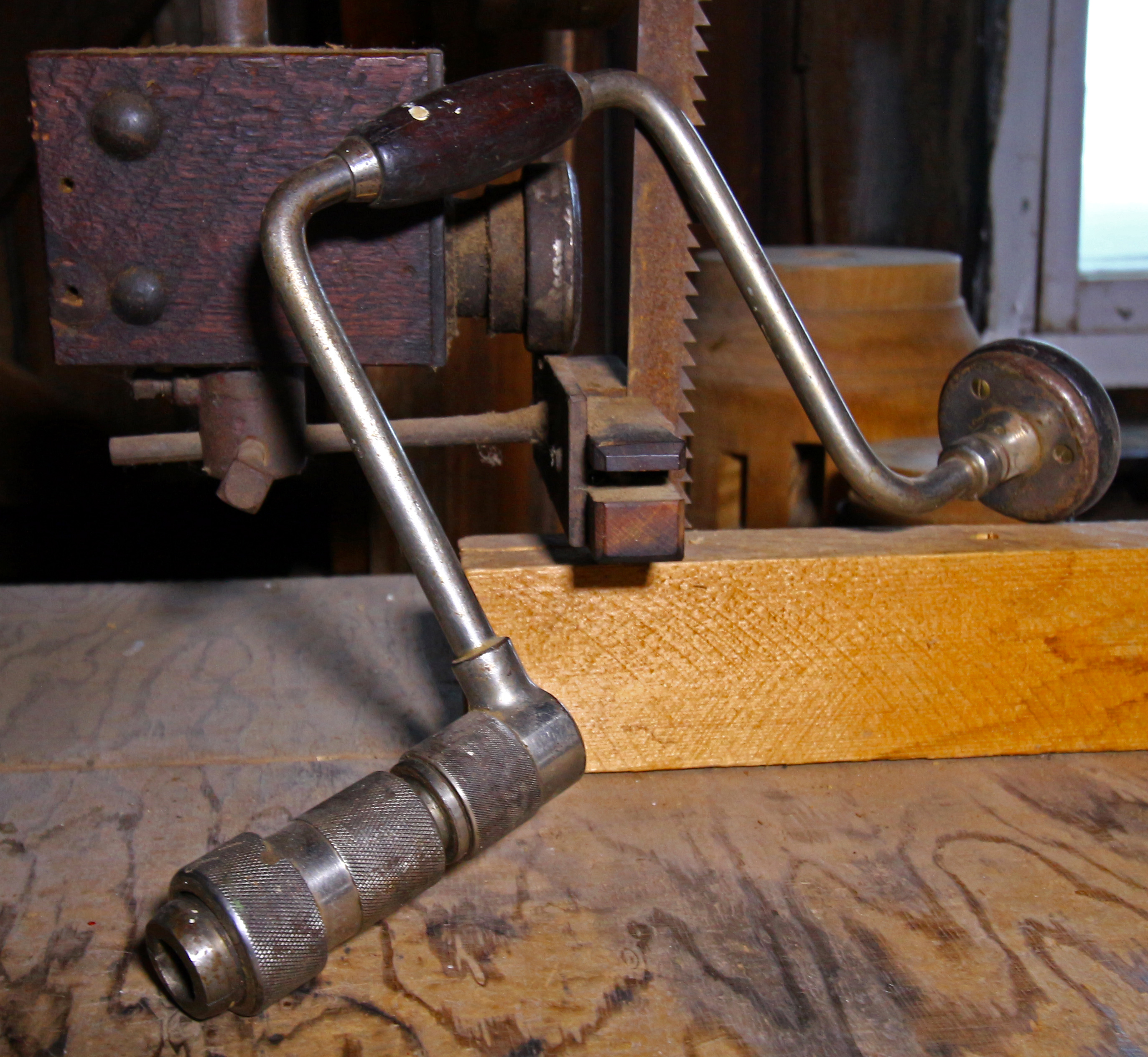 Close-up color photograph of a brace, made of metal with a wooden handle, set on a wooden workbench and leaning against a vintage band saw.