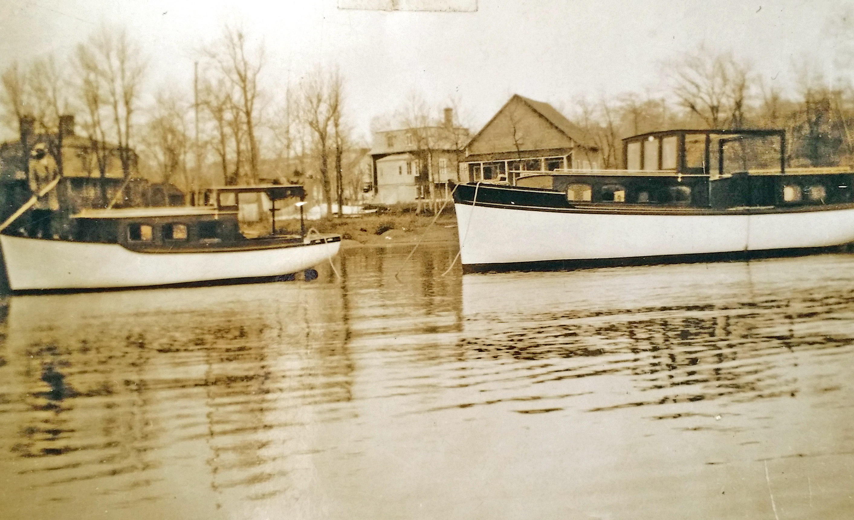Sepia photograph of two wooden boats, with cabin, anchored near the shore. Houses and trees without foliage are in the background.