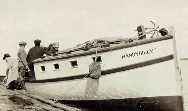 Black and white photograph of a yacht with the inscription Handy Billy, docked at a wooden wharf. A woman, two men and a boy get on board.