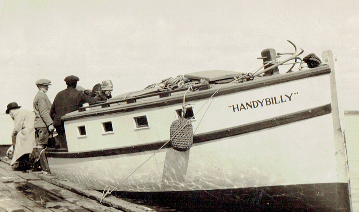 Black and white photograph of a yacht with the inscription "Handy Billy", docked at a wooden wharf. A woman, two men and a boy get on board.