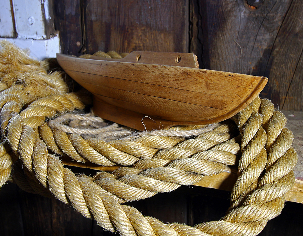 Color photograph of a wooden half hull, profile view. The object is placed on boat ropes.