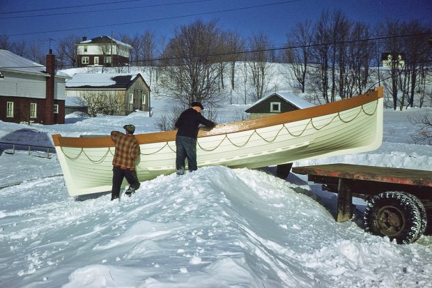 Color photograph where two men, one young and one elderly, slide a white boat on a pile of snow to install it on the rear platform of a truck.