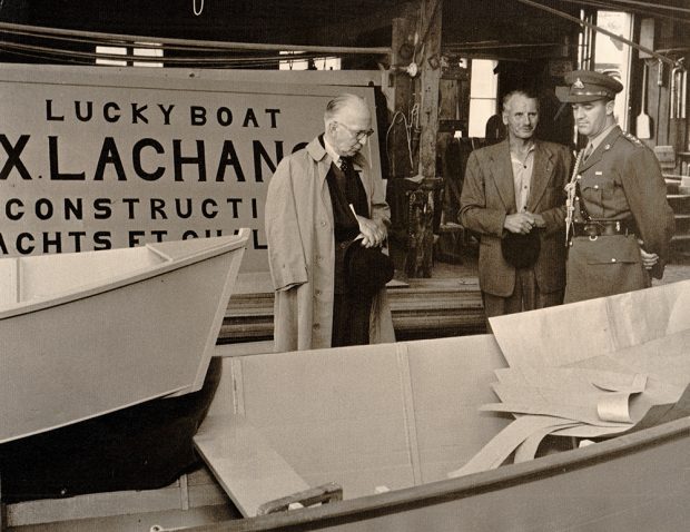 Black and white photograph where three men stand in a rowboat shop. A sign bearing the inscription Lucky boat. FX. Lachance. Construction de yachts et chaloupes is partially visible behind them. The men on the left (the Governor General) and on the right (a military) look at the boats. F-X Lachance is in the center, holding a hat.