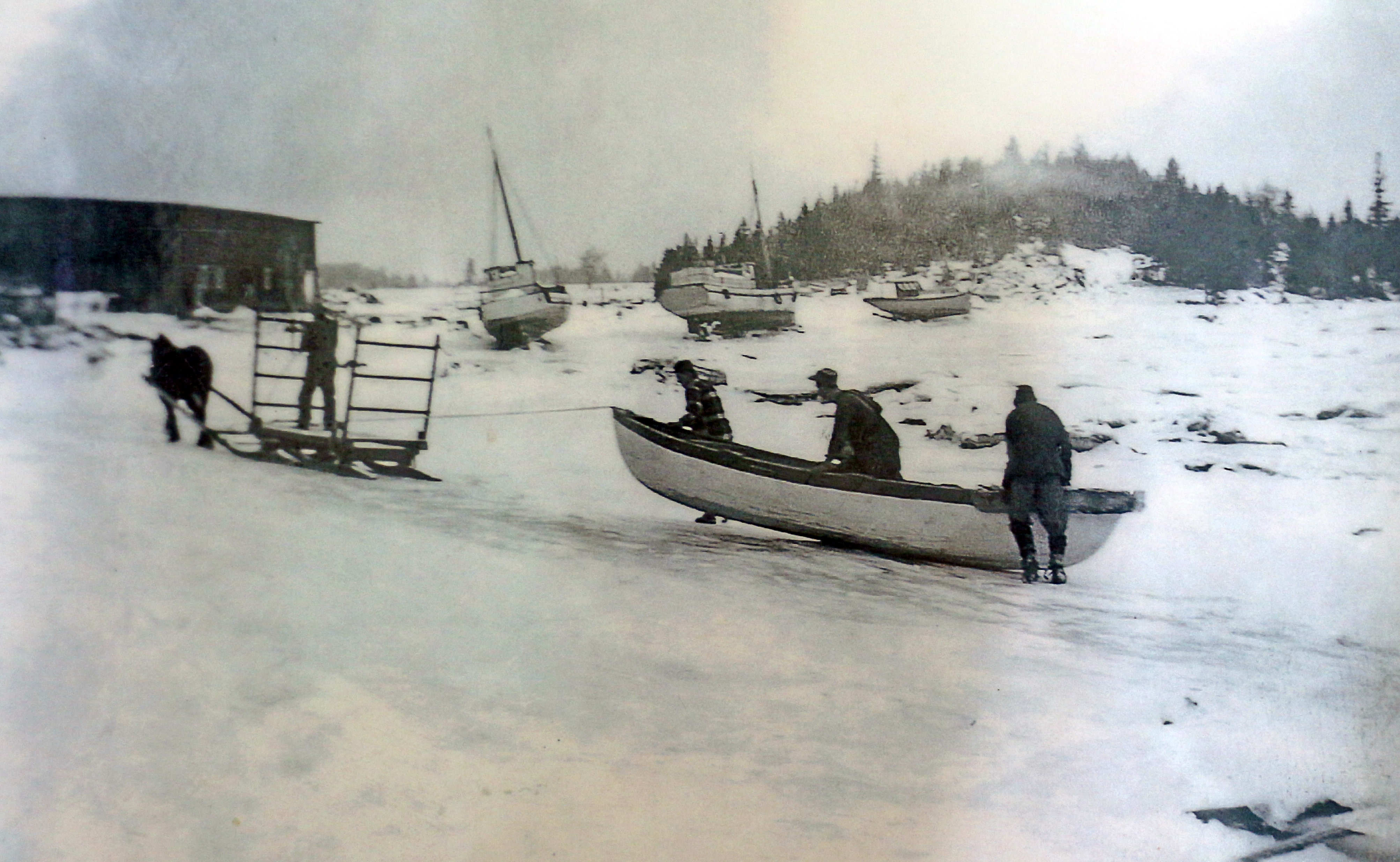 Black and white photograph where three men push a rowboat on the snowy shore. The boat is tied behind a sledge pulled by a horse on which stands a coachman. In the background are a building and three sailboats on the shore.