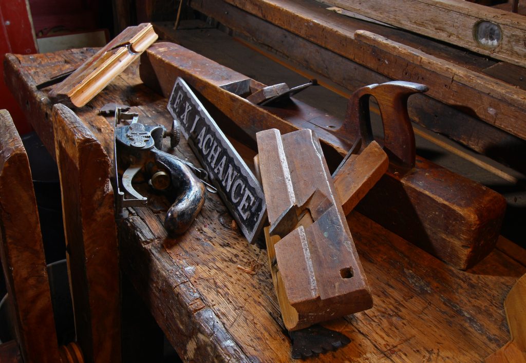 Current photography showing traditional carpentry tools, including a planer, a punch and a jig. The tools are placed on a workbench, next to a metal plate inscribed with 