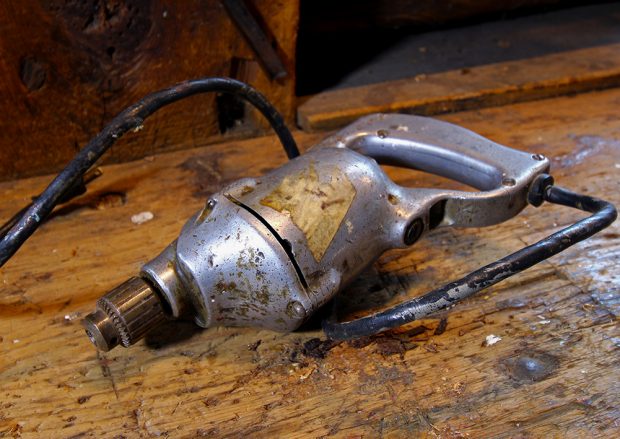 Close-up color photograph of a vintage electric drill, silver, set on a wooden workbench.