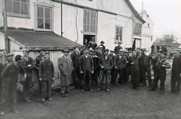 Black and white photograph of a group of about thirty workers posing in front of the side wall of a plank building, outside. The men are dressed in a working bitch or overalls. Many smoke and smile while looking at the camera. Others look towards François-Xavier Lachance, in the center. To his right is a man wearing a cloak, tie and hat.