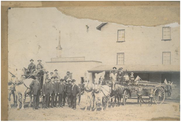 Men standing outside a stone mill with a decorated horse and carriage.