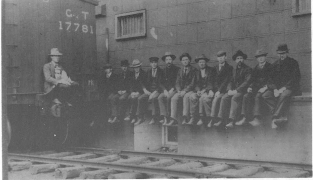 12 men sitting on a ledge by a train.